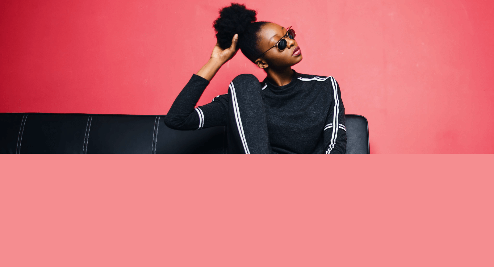 Modelo feminina negro com a mão no cabelo coque afro e óculos escuro fundo cor pêssego.